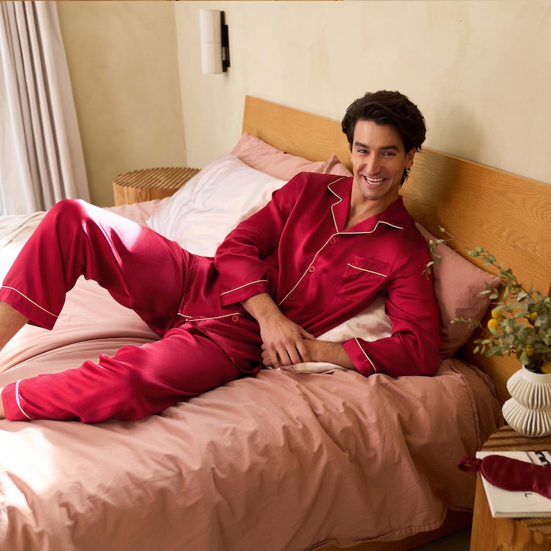 Op de afbeelding: Een persoon draagt een rode zijden pyjamaset met witte piping. De pyjama is langmouwig en heeft een knoopsluiting. De persoon ligt op een bed met roze lakens.