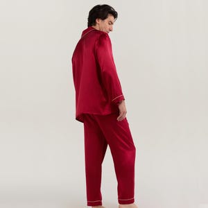 Op de afbeelding: Een rode zijden pyjamaset met een witte bies. Het bovenstuk heeft lange mouwen en een kraag. De broek heeft een rechte pijp.