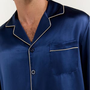 Op de afbeelding: Een donkerblauw overhemd met lange mouwen met een witte piping rond de kraag, manchetten en zak. Het overhemd heeft een knoopsluiting aan de voorkant en &eacute;&eacute;n zak op de linkerborst.