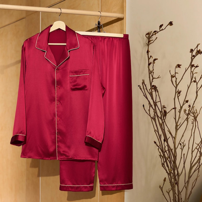 Op de afbeelding: Een rode satijnen pyjamaset met lange mouwen en broek. Het bovenstuk heeft een witte rand en een knoopsluiting. De broek heeft een witte rand aan de onderkant.