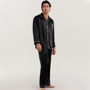 Op de afbeelding: Zwarte satijnen pyjama met witte piping. Het bovenstuk heeft een knoopsluiting en een kraag. De broek heeft een rechte pijp.