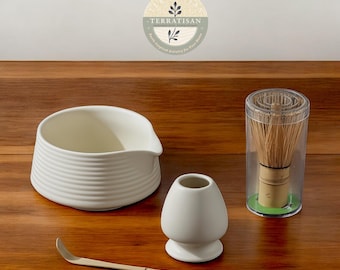 May include: A white ceramic matcha bowl, a white ceramic matcha whisk holder, a bamboo matcha whisk in a clear plastic container, and a gold metal matcha whisk. The items are arranged on a wooden surface. The text "TERRATISAN" is visible in the background.