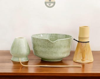 May include: A green ceramic matcha bowl, a green ceramic matcha whisk holder, a bamboo matcha whisk, and a bamboo matcha whisk holder on a wooden table.