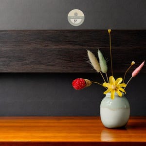 Puede incluir: Un pequeño jarrón de cerámica gris claro con un borde blanco se encuentra sobre una superficie de madera. El jarrón contiene un ramo de flores secas, incluyendo flores amarillas, bayas rojas y hierbas beige y verdes.