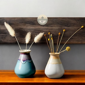 May include: Two ceramic vases with different colored glazes. The vase on the left is blue with a purple and white rim. The vase on the right is white with a yellow and brown rim. Both vases are filled with dried flowers.