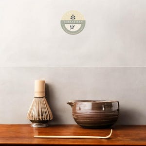 May include: A brown ceramic matcha bowl with a spout, a bamboo matcha whisk, and a wooden matcha scoop on a wooden table. The background is a light gray wall with a logo that says "Terratisan: Handcrafted Serenity For Your Home".