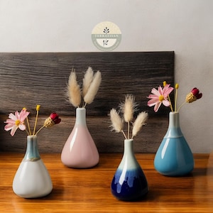 May include: Four small, ceramic vases in pastel colors, each holding dried flowers. The vases are in shades of white, pink, blue, and navy, with a glossy finish. The arrangement sits on a wooden surface, creating a minimalist aesthetic.