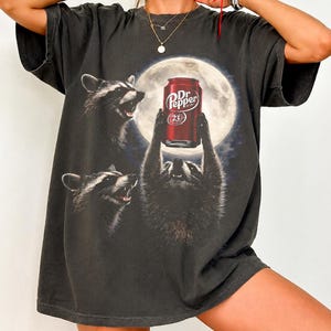 May include: A charcoal gray short-sleeve t-shirt featuring a graphic print of three raccoons under a full moon. One raccoon holds a red Dr. Pepper soda can above its head. The design includes the text Dr. Pepper and the number 23 on the can.
