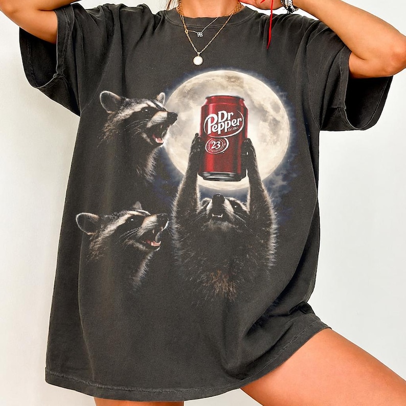 May include: A charcoal gray short-sleeve t-shirt featuring a graphic print of three raccoons under a full moon. One raccoon holds a red Dr. Pepper soda can above its head. The design includes the text Dr. Pepper and the number 23 on the can.