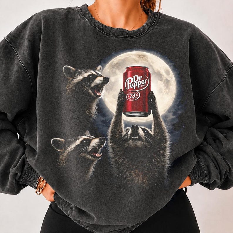 May include: A charcoal grey crewneck sweatshirt featuring a graphic print of three raccoons under a full moon. One raccoon holds a red Dr Pepper soda can aloft. The design includes the text Dr Pepper and the number 23 on the red aluminum beverage can.