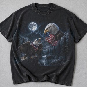 May include: A charcoal black short-sleeve t-shirt featuring a graphic print of two bald eagles in a night forest setting. One eagle holds an American flag in its beak, while the other wears a flag-patterned bandana. A full moon shines above.