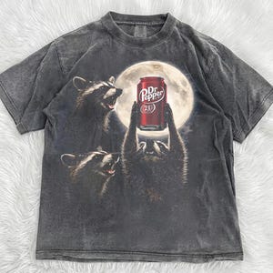 May include: A charcoal grey short-sleeve t-shirt featuring a graphic print of three raccoons under a full moon. One raccoon holds a red Dr Pepper soda can above its head. The shirt has a faded, washed-out appearance with a crew neckline.