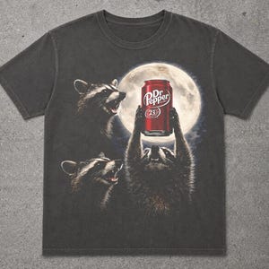 May include: A charcoal gray short-sleeve t-shirt featuring a graphic print of three raccoons under a full moon. One raccoon holds a red Dr. Pepper soda can above its head. The design includes the text Dr. Pepper and the number 23 on the can.