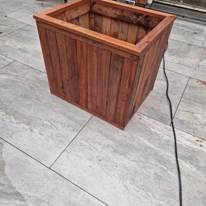 May include: A rectangular wooden planter box with a natural wood finish. The planter is empty and sits on a gray tiled floor.