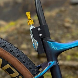 以下が含まれることがあります： 黒い自転車のシートポストにCO2インフレーターが取り付けられています。インフレーターは金色のヘッドで、銀色のCO2カートリッジに接続されています。自転車のフレームはダークブルーで、虹色のハイライトがあり、茶色のタイヤが見えます。