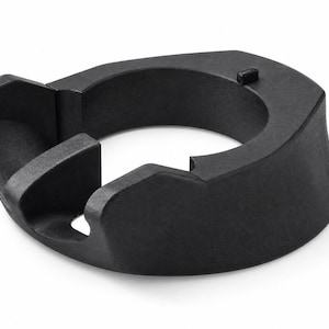 Puede incluir: Un soporte circular de plástico negro mate con una abertura central y un borde contorneado con una pequeña pestaña que sobresale. El objeto tiene una base ancha y estable y una sección recortada en un lado, con un acabado uniforme.