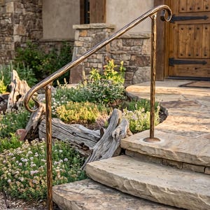 May include: A metal handrail with a textured bronze finish and scrolled ends, mounted on stone steps. The railing features a single vertical support post and an angled top bar, providing a sturdy grip for navigating the outdoor stone staircase.