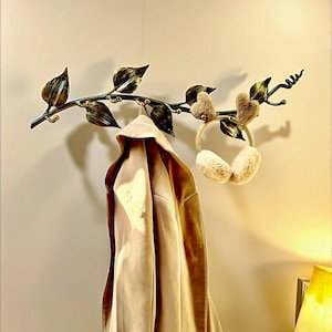 Puede incluir: Un gancho de pared decorativo de metal con forma de rama con hojas, que sostiene un abrigo beige y orejeras esponjosas. El metal tiene un acabado en bronce y oro. El abrigo es de color beige claro.