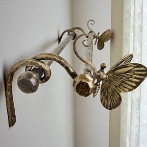Puede incluir: Barra de cortina de estilo antiguo con detalles decorativos de mariposas. La barra de metal tiene un acabado en bronce. Los detalles de mariposa añaden un toque de elegancia. La barra está montada en una pared blanca.