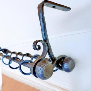 Może przedstawiać: Kuty żelazny drążek do zasłon z dekoracyjnymi zawijasami i detalami pierścieni. Drążek ma ciemne, teksturowane wykończenie i jest przeznaczony do montażu na ścianie. Projekt obejmuje wspornik montażowy i zakończenia.