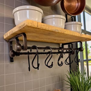 May include: A rustic wooden shelf with black metal brackets and hooks. Three white ceramic bowls sit on the shelf. Copper pots hang above, and a potted plant is in the foreground. Kitchen decor.