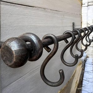 Può includere: Un bastone per tende in ferro battuto con terminali decorativi e ganci a forma di S. Il bastone è montato su una parete di legno grigio chiaro. Il metallo ha una finitura scura e invecchiata, che suggerisce uno stile rustico o vintage. I ganci sono distanziati uniformemente.