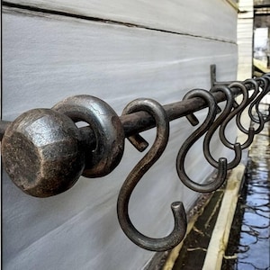 May include: A wrought iron curtain rail with decorative end pieces and S-shaped hooks. The rail is mounted on a light grey wall. The metal has a dark, aged finish, suggesting a rustic or vintage style. The hooks are evenly spaced.