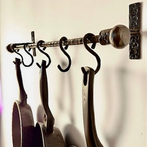 Puede incluir: Un estante de cocina de hierro forjado con seis ganchos en forma de S, que sostiene tres sartenes oscuras. El estante está montado en una pared blanca. El estante tiene una pieza final decorativa y un soporte de pared a juego.