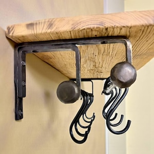 May include: A rustic wooden shelf with dark metal brackets and decorative knobs. Below the shelf hang several black metal S-hooks, perfect for hanging items. The shelf is mounted on a light-colored wall.