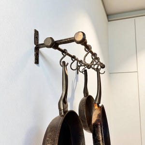 Könnte beinhalten: Ein schmiedeeiserner Topfhalter mit hängenden Pfannen. Der Halter ist an einer weißen Wand montiert und verfügt über eine horizontale Stange mit dekorativen Haken. Drei dunkle Metallpfannen mit langen Griffen hängen an den Haken.