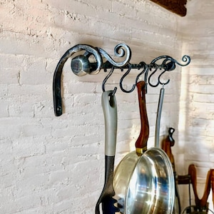 Könnte beinhalten: Ein dekorativer Topfhalter aus Schmiedeeisen mit sechs Haken, montiert an einer weißen Backsteinmauer. Ein schwarzer Nudellöffel, eine Edelstahlpfanne und andere Küchenutensilien hängen an den Haken. Über dem Gestell befindet sich ein Holzbalken.