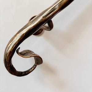 May include: A handrail with a textured, dark bronze finish featuring a curved end that terminates in a decorative leaf-like scroll. The metal bar has a hammered appearance and is secured to the wall by a circular mounting bracket.