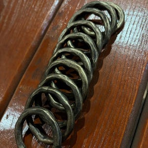 Könnte beinhalten: Eine Ansammlung antiker, bronzefarbener Ringe, jeweils mit einer strukturierten, unregelmäßigen Oberfläche. Die Ringe sind in einer leicht gebogenen Linie angeordnet, was darauf hindeutet, dass sie verbunden sein könnten. Sie liegen auf einer Holzoberfläche.
