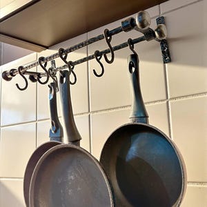 May include: A wrought iron pot rack with three hanging pans. The rack is mounted on a white tiled wall and features decorative S-hooks. The pans are dark grey with long handles, ready for cooking.