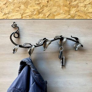 Puede incluir: Un perchero de metal con forma de vid con hojas. El perchero tiene tres ganchos y está montado en una pared de color marrón claro. Una chaqueta azul está colgada en uno de los ganchos y un juego de llaves está colgado en otro gancho.
