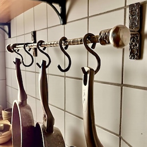 Puede incluir: Un estante para ollas de hierro forjado con cinco ganchos en forma de S, que sostiene tres sartenes oscuras. El estante está montado en una pared alicatada con una estantería de madera encima. El estante tiene piezas decorativas en los extremos y una barra texturizada.