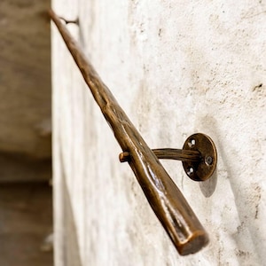 Puede incluir: Una barandilla de color bronce con un diseño orgánico texturizado, montada en una pared clara. La barandilla está soportada por soportes circulares con fijaciones de tornillo, creando una estética rústica y elegante. La barandilla parece ser de metal.