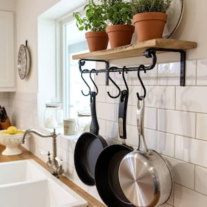 May include: A kitchen scene featuring a wooden shelf with three potted herbs and a decorative plate. Below, a black metal rack holds three pans. The kitchen has white subway tile and a wooden countertop.