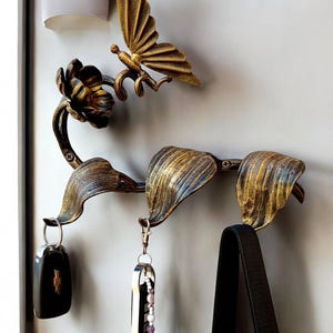 May include: Ornate, gold-colored metal wall hook with a butterfly and floral design. The hook holds keys and a black bag. The design includes a butterfly, flower, and leaf-shaped hooks, adding a decorative touch to any space.