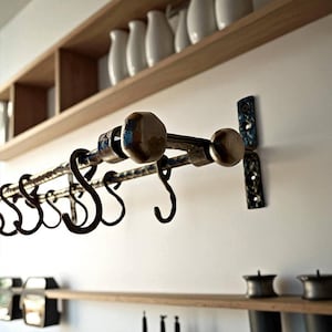 Puede incluir: Un estante de cocina de hierro forjado con ganchos en forma de S decorativos y remates. El estante está montado en una pared blanca con una estantería de madera encima. El metal tiene un aspecto envejecido con detalles azules.