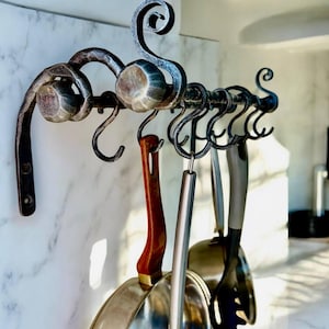 Puede incluir: Un estante de cocina de hierro forjado con ganchos decorativos en forma de cisne. El estante sostiene varios utensilios de cocina y una sartén de acero inoxidable con mango de madera. El estante está montado en una pared de mármol blanco.