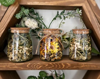 May include: Three glass jars with cork lids filled with dried flowers and herbs. The jars are arranged on a wooden shelf with a rustic brown finish.
