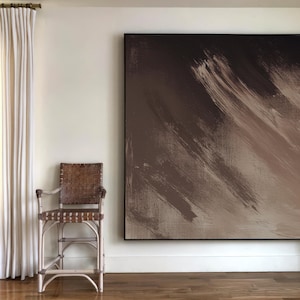 May include: A white wicker chair with a brown woven seat and back sits in front of a large abstract painting with brown and beige brushstrokes. The painting is framed in black.