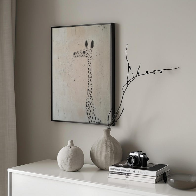 May include: A black framed print of a giraffe with black spots on a light pink background. The print is hanging on a light gray wall above a white dresser. There are two gray ceramic vases on the dresser, one with a round shape and the other with a bulbous shape. A black camera and a stack of books are also on the dresser.