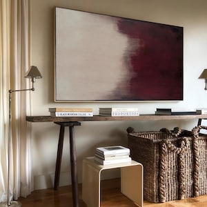 May include: A rustic wooden console table with a large abstract painting featuring a gradient from white to burgundy. The table is decorated with books and a woven basket.