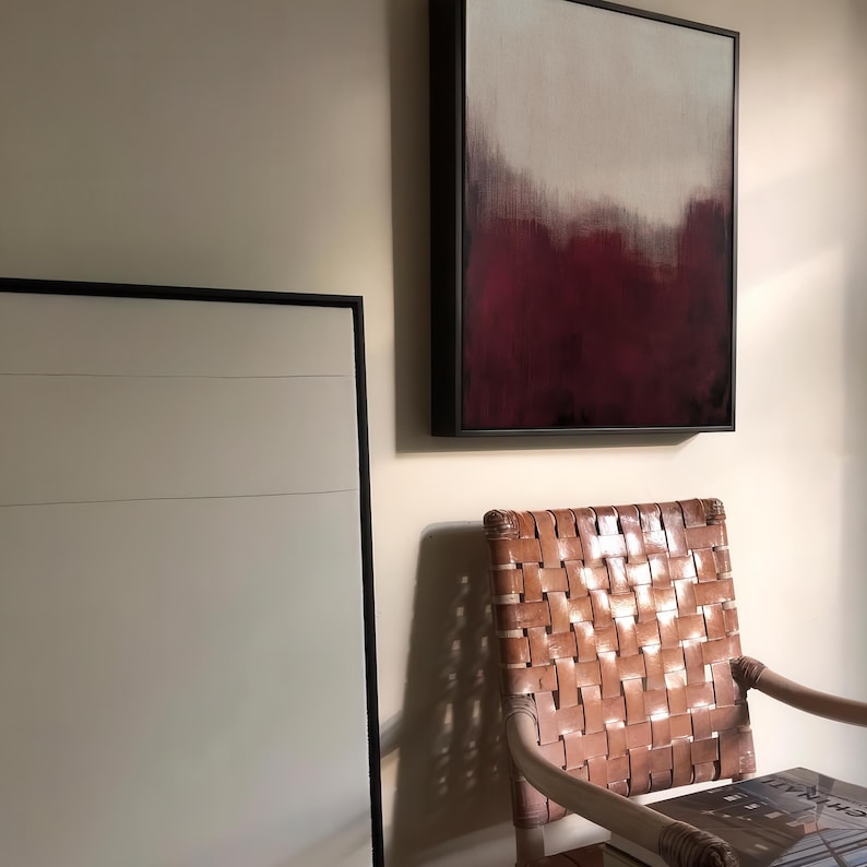 May include: A brown leather chair with a woven back and seat. The chair is in front of a white wall with two framed paintings. One painting is a landscape with a red and white colour scheme.