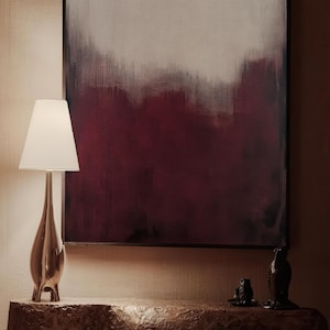May include: A modern, silver-toned table lamp with a white lampshade sits on a wooden table in front of a framed abstract painting with a gradient of red and grey.