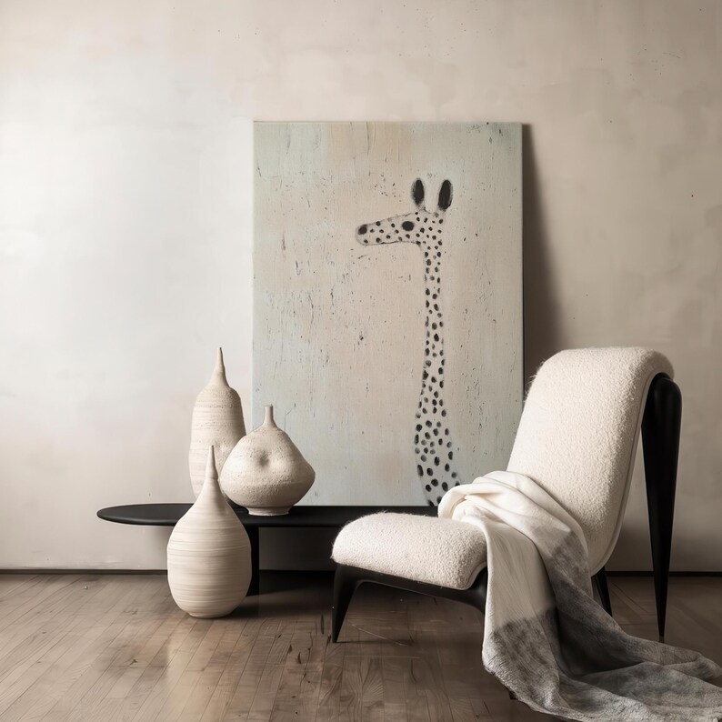 May include: A white chair with a black frame and a white throw blanket sits in front of a painting of a giraffe with black spots on a light pink background. Three white ceramic vases are on a black table in front of the chair.