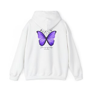 Könnte beinhalten: Weißer Hoodie mit einem lila Schmetterlingsmotiv auf der Rückseite. Der Schmetterling hat schwarze Umrisse und ist von kleineren schwarzen Schmetterlingen umgeben. Der Text "Glaube an deine Flügel, auch wenn du sie nicht sehen kannst" ist unter dem Schmetterling gedruckt.