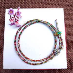 Puede incluir: Un collar trenzado multicolor con hilos rojos, verdes, amarillos y plateados, sobre una superficie blanca. El collar tiene un cierre plateado y un pequeño acento turquesa. Una ramita de flores moradas está presente.